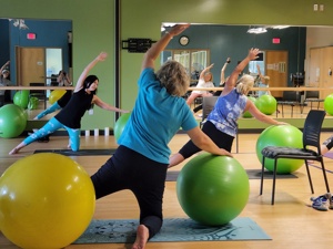 people in a fitness room kneeling on yoga mats and stretching their arm overhead and extending one leg while balancing on a big fitness ball. Following an instructor in front of the room and facing mirrors that line the wall.